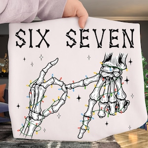 May include: White tote bag featuring a skeletal hand gesture spelling out "SIX SEVEN" in black gothic lettering. The hands are decorated with colorful Christmas lights and surrounded by star-shaped accents.