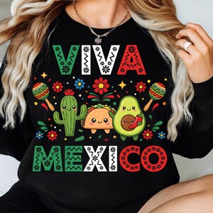 Op de afbeelding: Zwarte sweatshirt met de tekst "VIVA MEXICO" in groen, wit en rood. Het ontwerp bevat een cactus, een taco en een avocado met lachende gezichten, maracas en kleurrijke bloemen.