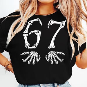 May include: Black t-shirt featuring a white skeleton design. The design includes the number "67" in a bone-like font, with two skeleton hands below. The shirt is a casual, Halloween-themed apparel item.