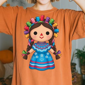 K&ouml;nnte beinhalten: Orangefarbenes T-Shirt mit einer farbenfrohen Illustration einer traditionellen mexikanischen Puppe. Die Puppe hat dunkles Haar, ein blaues Kleid mit wei&szlig;en floralen Details und ein lebhaftes Stirnband aus B&auml;ndern. Die Z&ouml;pfe der Puppe sind mit bunten B&auml;ndern verziert.