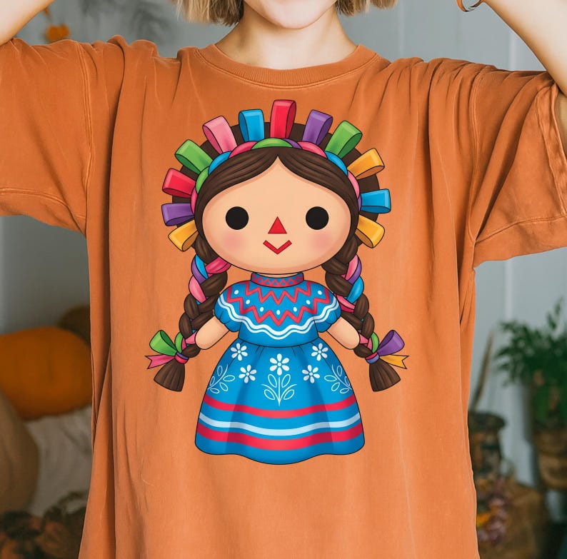 K&ouml;nnte beinhalten: Orangefarbenes T-Shirt mit einer farbenfrohen Illustration einer traditionellen mexikanischen Puppe. Die Puppe hat dunkles Haar, ein blaues Kleid mit wei&szlig;en floralen Details und ein lebhaftes Stirnband aus B&auml;ndern. Die Z&ouml;pfe der Puppe sind mit bunten B&auml;ndern verziert.