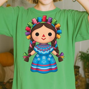 K&ouml;nnte beinhalten: Gr&uuml;nes T-Shirt mit einer farbenfrohen Illustration einer traditionellen mexikanischen Puppe. Die Puppe hat dunkles Haar, geflochtene Z&ouml;pfe und ein blaues Kleid mit floralen und gestreiften Details. Der Kopf der Puppe ist mit einem Stirnband aus bunten B&auml;ndern geschm&uuml;ckt.