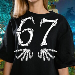 May include: Black t-shirt with a white skeleton bone design. The design features the number "67" in a bone font, with two skeletal hands below. The shirt is a casual, crew neck style.