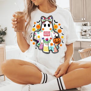 May include: White t-shirt featuring a ghost design with a colorful floral and Day of the Dead-inspired pattern. The ghost holds a jack-o'-lantern and is surrounded by bats, flowers, and spiderwebs. The shirt has a black lace trim.