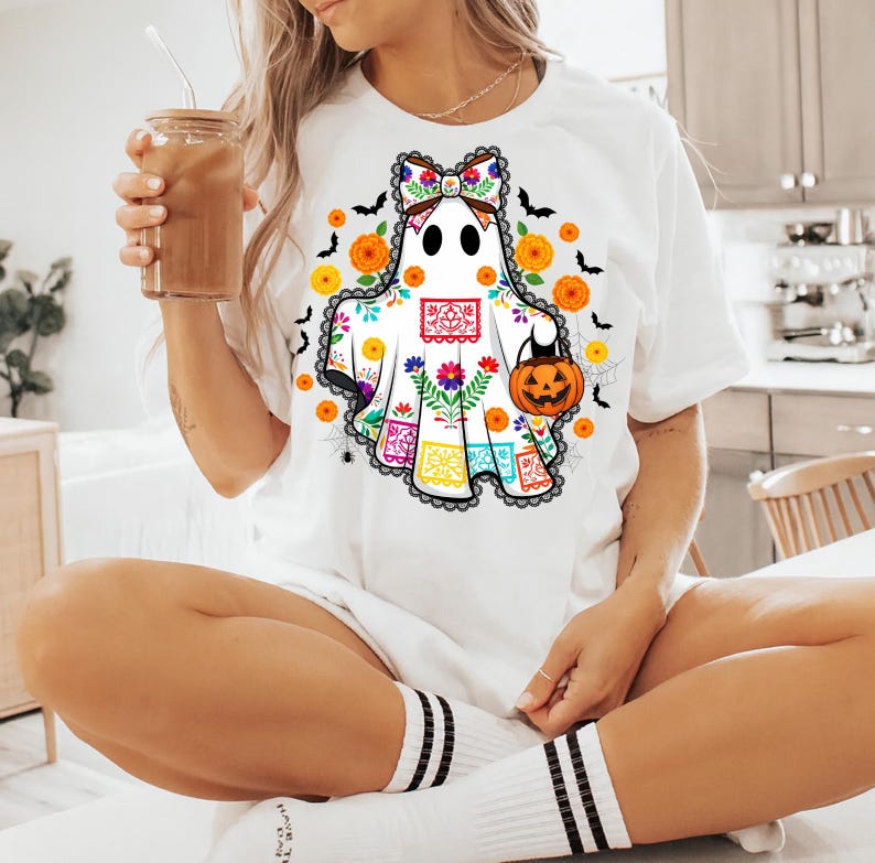 May include: White t-shirt featuring a ghost design with a colorful floral and Day of the Dead-inspired pattern. The ghost holds a jack-o'-lantern and is surrounded by bats, flowers, and spiderwebs. The shirt has a black lace trim.