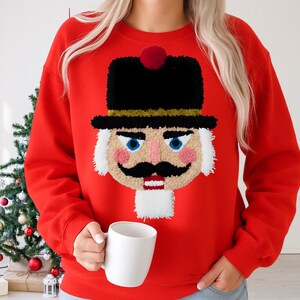 Puede incluir: Sudadera roja con un diseño bordado de cascanueces. El cascanueces tiene un sombrero negro con un pompón rojo, un bigote negro y una barba blanca. Un diseño festivo navideño.