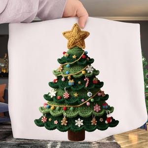 May include: A green crocheted Christmas tree pillow with a gold star topper. The tree is decorated with colorful ornaments, gingerbread men, snowflakes, and gift boxes. The pillow is held up by a person's hand.