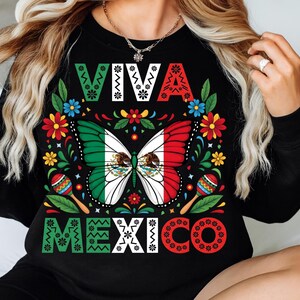 Op de afbeelding: Zwart shirt met lange mouwen en een kleurrijk ontwerp. Het ontwerp toont de woorden "VIVA MEXICO" in wit, groen en rood. Een vlinder met de kleuren van de Mexicaanse vlag staat in het midden, omringd door bloemen en maracas.