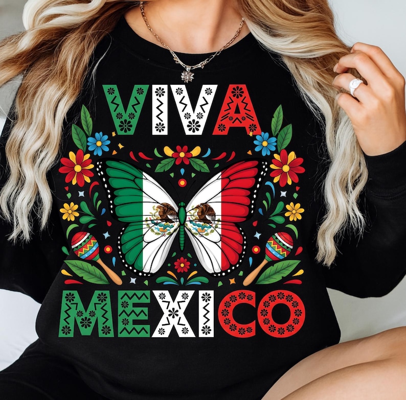 Op de afbeelding: Zwart shirt met lange mouwen en een kleurrijk ontwerp. Het ontwerp toont de woorden "VIVA MEXICO" in wit, groen en rood. Een vlinder met de kleuren van de Mexicaanse vlag staat in het midden, omringd door bloemen en maracas.