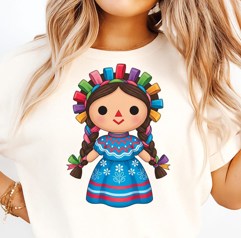 May include: A graphic of a traditional Mexican doll, wearing a blue dress with white floral details and a colorful headband. The doll has brown braids and a smiling face. The design is printed on a cream-colored t-shirt.