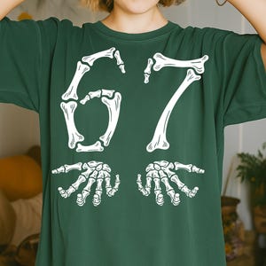 May include: Dark green t-shirt with a white skeleton-themed design. The design features the number "67" in a skeletal font, with two skeletal hands below. The shirt is made of a soft, comfortable material.