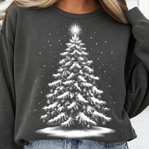 May include: Dark gray sweatshirt featuring a white Christmas tree design. The tree is decorated with ornaments and lights, and topped with a star. Snowflakes are falling around the tree, creating a festive winter scene.