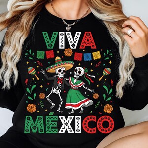 Op de afbeelding: Zwart shirt met lange mouwen met een kleurrijke afbeelding ter ere van D&iacute;a de Muertos. Het ontwerp toont dansende skeletten, maracas en de woorden "VIVA MEXICO" in feestelijke letters. Het shirt is versierd met bloemen en banners.