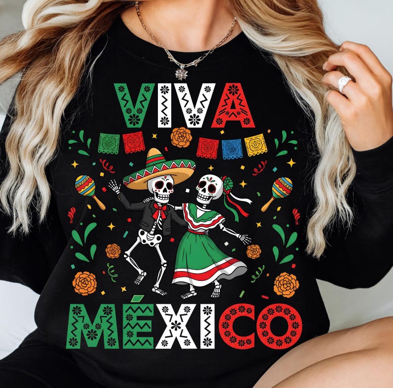 Op de afbeelding: Zwart shirt met lange mouwen met een kleurrijke afbeelding ter ere van D&iacute;a de Muertos. Het ontwerp toont dansende skeletten, maracas en de woorden "VIVA MEXICO" in feestelijke letters. Het shirt is versierd met bloemen en banners.