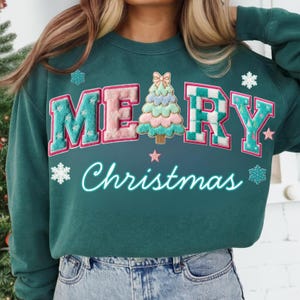 May include: Teal green sweatshirt with the words "MERRY Christmas" in a playful, colorful design. The letters are in a varsity style with a Christmas tree graphic. Snowflake and star accents complete the festive look.