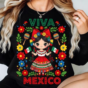 May include: Black sweatshirt with a colorful graphic featuring a cartoon girl in a traditional Mexican dress, holding Mexican flags. The design includes the words "VIVA MEXICO" and decorative flowers and maracas.