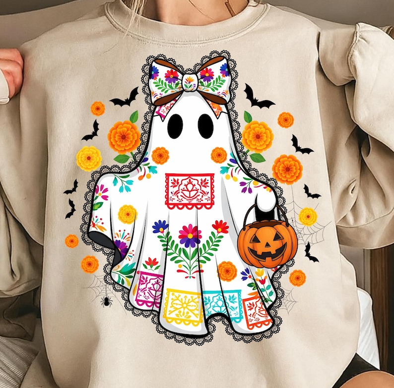 May include: A beige sweatshirt featuring a white ghost with a colorful floral design and a pumpkin-shaped bag. The ghost has a decorative bow and is surrounded by orange flowers, bats, and spiderwebs, perfect for Halloween.