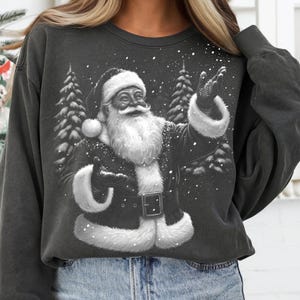 May include: Dark gray sweatshirt featuring a black and white illustration of Santa Claus. Santa is depicted with a long white beard, a black coat with white fur trim, and a Santa hat. The background includes snowy trees and falling snow.