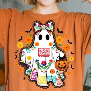 May include: Orange t-shirt featuring a white ghost with a colorful floral design and a jack-o'-lantern. The ghost has a bow and is surrounded by bats, flowers, and a spiderweb, perfect for Halloween or Day of the Dead celebrations.