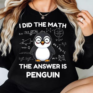 Op de afbeelding: Zwart sweatshirt met de tekst "I DID THE MATH THE ANSWER IS PENGUIN". Het ontwerp toont een cartoonpinguïn met een bril, omringd door wiskundige formules. De pinguïn heeft een witte buik en een lachend gezicht.