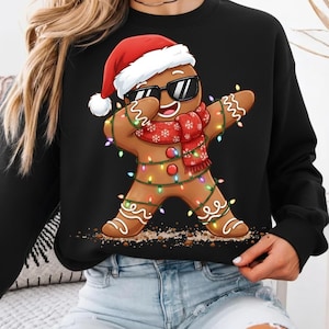 May include: Black sweatshirt featuring a gingerbread man wearing sunglasses and a Santa hat. The gingerbread man is dabbing and wearing a red scarf with snowflakes and is decorated with Christmas lights.