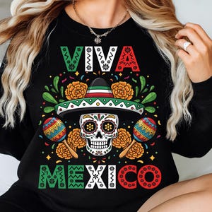Op de afbeelding: Zwarte sweatshirt met een kleurrijk "Viva Mexico" ontwerp. De afbeelding bevat een suikerschedel met een sombrero, maracas en bloemaccenten in groen, rood en geel. De teksten "VIVA" en "MEXICO" zijn ook aanwezig.