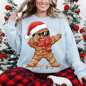 May include: Light blue sweatshirt featuring a gingerbread man wearing sunglasses, a Santa hat, and a red scarf. The gingerbread man is decorated with Christmas lights and is in a dabbing pose. The sweatshirt is part of a Christmas-themed collection.