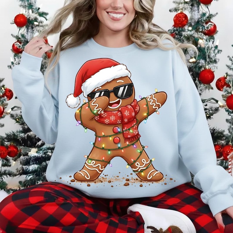 May include: Light blue sweatshirt featuring a gingerbread man wearing sunglasses, a Santa hat, and a red scarf. The gingerbread man is decorated with Christmas lights and is in a dabbing pose. The sweatshirt is part of a Christmas-themed collection.