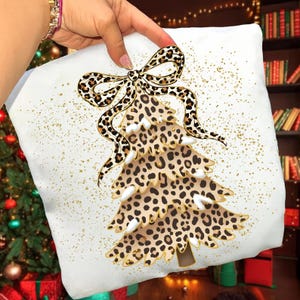 Puede incluir: Cojín cuadrado blanco con un diseño de árbol de Navidad con estampado de leopardo. El árbol está adornado con un lazo a juego con estampado de leopardo. Brillo dorado acentúa el fondo. El cojín es sostenido por una persona.