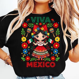 May include: Black t-shirt featuring a colorful illustration of a girl in traditional Mexican attire. The design includes the words "VIVA MEXICO" and decorative flowers, maracas, and the Mexican flag. The girl is wearing a red dress and holding two Mexican flags.