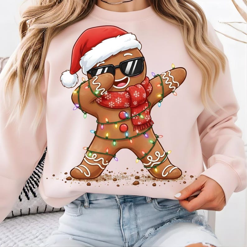 May include: A light pink sweatshirt featuring a gingerbread man illustration. The gingerbread man is wearing sunglasses, a Santa hat, and a red scarf. He is decorated with colorful Christmas lights and is doing the dab.