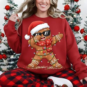 May include: Red sweatshirt featuring a dabbing gingerbread man wearing sunglasses, a Santa hat, and a scarf. The gingerbread man is decorated with Christmas lights. The person in the image is wearing red and black plaid pajama pants.