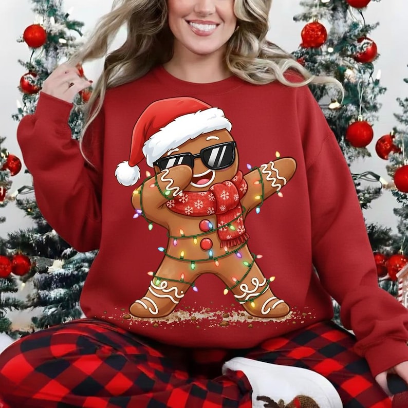 May include: Red sweatshirt featuring a dabbing gingerbread man wearing sunglasses, a Santa hat, and a scarf. The gingerbread man is decorated with Christmas lights. The person in the image is wearing red and black plaid pajama pants.