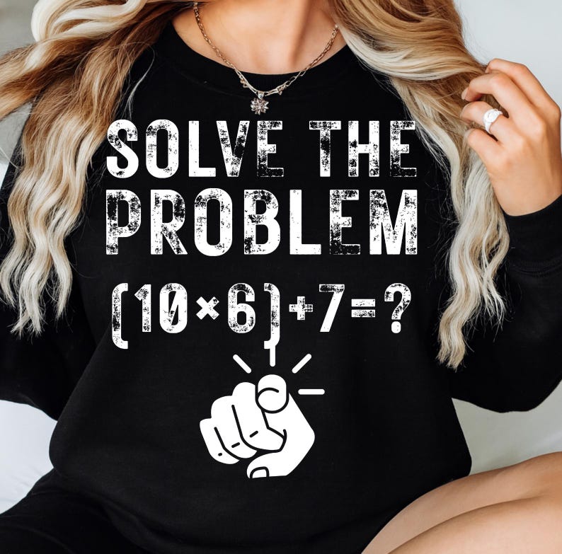 May include: Black sweatshirt with the text "SOLVE THE PROBLEM" in white, distressed lettering. Below the text is a math problem: (10*6)+7=?, with a pointing hand graphic. The sweatshirt is a casual style.