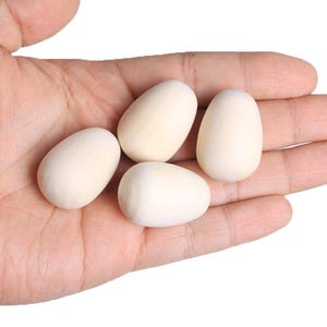 May include: Four smooth, white wooden eggs in a hand. The eggs are unpainted and ready for crafting.