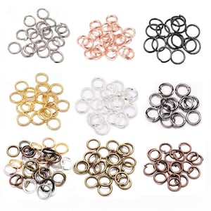 May include: A collection of jump rings in various metallic finishes, including silver, rose gold, black, gold, and bronze. These small, circular rings are used for jewelry making and crafting. The jump rings are arranged in separate piles.