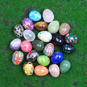 Puede incluir: Una colección de 20 huevos de piedras preciosas pulidas en varios colores y patrones, incluyendo verde, azul, rojo, blanco y marrón. Los huevos están dispuestos en un semicírculo sobre una superficie verde.