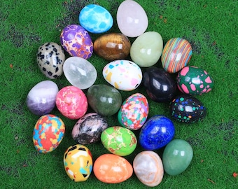 Puede incluir: Una colección de 20 huevos de piedras preciosas pulidas en varios colores y patrones, incluyendo verde, azul, rojo, blanco y marrón. Los huevos están dispuestos en un semicírculo sobre una superficie verde.