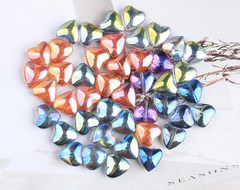 Op de afbeelding: Een verzameling iriserende hartvormige kralen in tinten oranje, paars, blauw en groen. De kralen zijn in een cluster gerangschikt, waardoor ze licht reflecteren en een glinsterend effect creëren. Deze decoratieve kralen zijn ideaal voor knutselen en sieraden maken.
