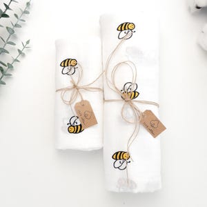 Può includere: Due fasce in mussola bianca con api ricamate e un'etichetta marrone che dice "Little Cotton Clouds". Le fasce sono legate con uno spago.