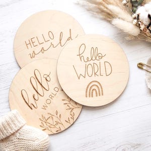 Può includere: Tre targhe rotonde in legno con le parole "Hello World" incise. Una targa presenta un disegno arcobaleno, e un'altra un'illustrazione botanica. Le targhe sono di colore marrone chiaro.