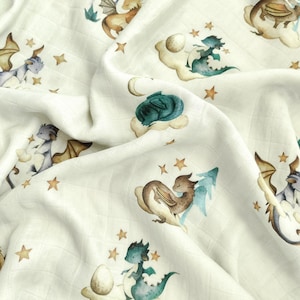 Op de afbeelding: Crèmekleurige stof met een herhaald patroon van aquarel draken, wolken, sterren en manen. De draken zijn in tinten blauw, bruin en grijs, met gouden accenten. De stof heeft een zachte, getextureerde uitstraling.