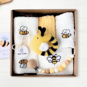 Op de afbeelding: Een cadeaudoos gevuld met babyartikelen met een geel en zwart bijenmotief. De doos bevat een gehaakte bijenrammelaar, een houten bijtring, een kralenketting en vier witte katoenen doeken met geborduurde bijen.