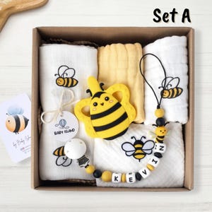 Può includere: Un set regalo a tema ape. Il set include un mordedor di silicone a forma di ape giallo e nero, un panno di mussola bianco con un disegno di ape, un panno di mussola giallo, un panno di mussola bianco con un disegno di ape e il nome "Kevin" scritto in perline bianche e nere, e una collana di perline nere e gialle con un ciondolo a forma di ape.