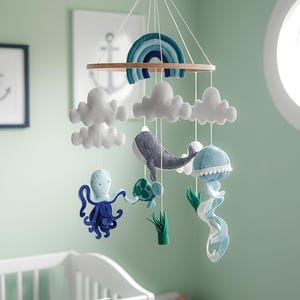 Può includere: Mobile per neonati con un anello di legno, un arcobaleno in feltro e nuvole bianche. Include creature marine come una balena, un polpo, una tartaruga, una medusa e alghe nei toni del blu, verde e bianco. Appeso sopra una culla bianca.