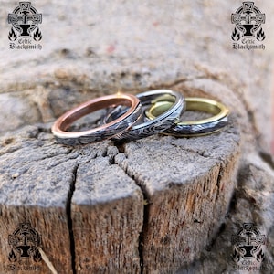 May include: Three rings made of hammered metal with a wood grain pattern. The rings are stacked on top of each other. The top ring is gold, the middle ring is silver, and the bottom ring is copper.