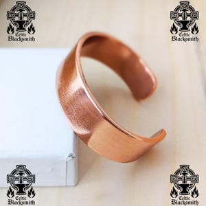 Pode incluir: Uma pulseira de punho de cobre com acabamento escovado. A pulseira aberta tem um design curvo e repousa sobre uma superfície branca. O logotipo Celtic Blacksmith é visível nos cantos da imagem.