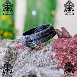 Pode incluir: Um anel de aço Damasco com um padrão único de metal em camadas. O anel é cinza escuro com linhas cinza mais claras. O logotipo Celtic Blacksmith é visível no fundo. O anel é exibido em uma superfície texturizada marrom-avermelhada.