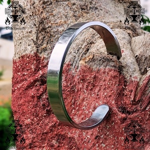 May include: A silver-coloured, open-ended cuff bracelet. The bracelet has a flat, rectangular profile and a smooth, polished surface. The bracelet is displayed against a textured, reddish-brown background. The text "Celtic Blacksmith" is visible.