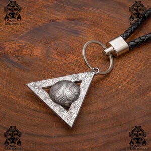 Könnte beinhalten: Ein silberner dreieckiger Schlüsselanhänger mit einer runden, gemusterten Metallscheibe in der Mitte. Der Schlüsselanhänger ist an einer schwarzen Lederschnur mit einem silbernen Ring befestigt.